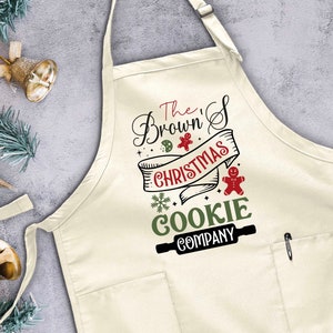 Puede incluir: Un delantal blanco con un bolsillo y un gr&aacute;fico de rodillo de amasar marr&oacute;n. El texto en el delantal dice "The Brown's Christmas Cookie Company".