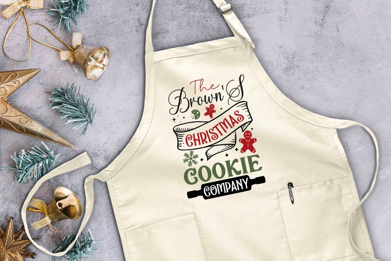 Puede incluir: Un delantal blanco con un bolsillo y un gr&aacute;fico de rodillo de amasar marr&oacute;n. El texto en el delantal dice "The Brown's Christmas Cookie Company".