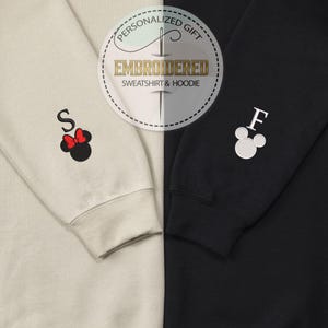 May include: Two sweatshirts, one beige and one black, with embroidered Mickey Mouse ears and letters. The beige sweatshirt has a red bow and the letter "S" embroidered on the sleeve. The black sweatshirt has the letter "F" embroidered on the sleeve. The sweatshirts are personalized gifts.