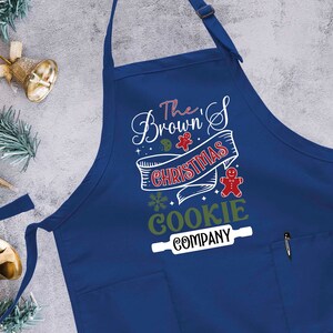 Puede incluir: Delantal azul con tem&aacute;tica navide&ntilde;a con el texto "The Brown's Christmas Cookie Company" en blanco con acentos verdes y rojos. Un gr&aacute;fico de galleta de hombre de jengibre est&aacute; en el delantal.