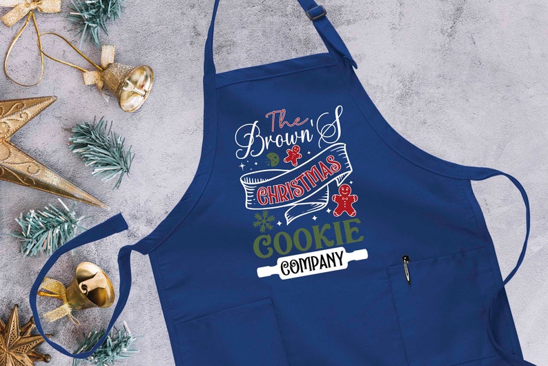Puede incluir: Delantal azul con tem&aacute;tica navide&ntilde;a con el texto "The Brown's Christmas Cookie Company" en blanco con acentos verdes y rojos. Un gr&aacute;fico de galleta de hombre de jengibre est&aacute; en el delantal.