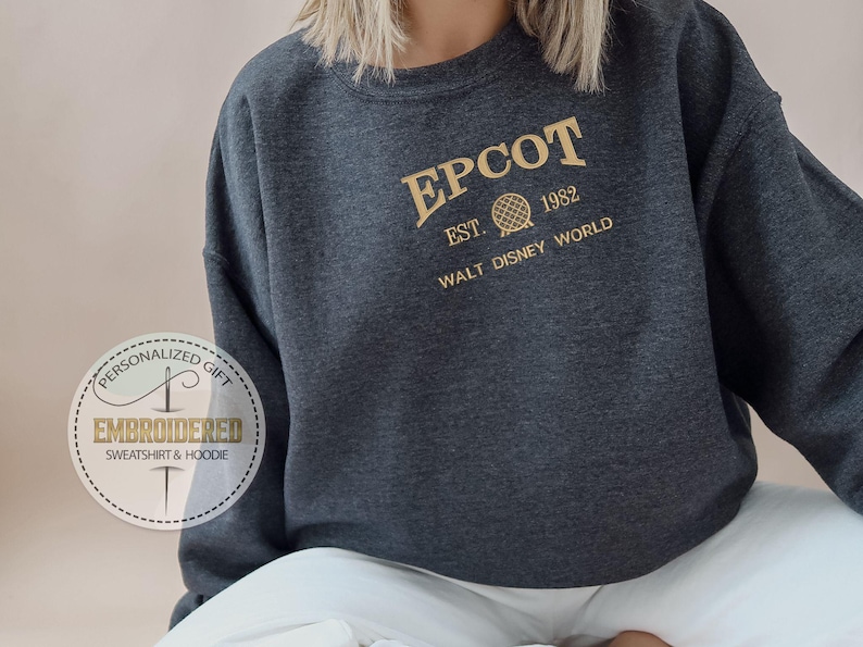 Puede incluir: Sudadera EPCOT gris oscuro con letras doradas. El texto incluye "EPCOT", "EST. 1982" y "WALT DISNEY WORLD". La sudadera tiene cuello redondo y mangas largas. A la izquierda hay una pegatina redonda con el texto "EMBROIDERED SWEATSHIRT & HOODIE".