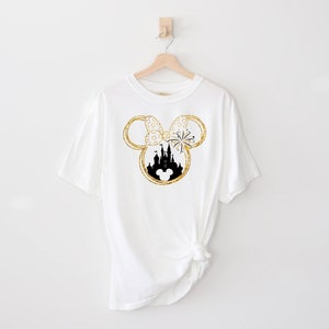 May include: White t-shirt with a gold glitter Mickey Mouse ears design featuring a black silhouette of Cinderella's castle inside the ears.