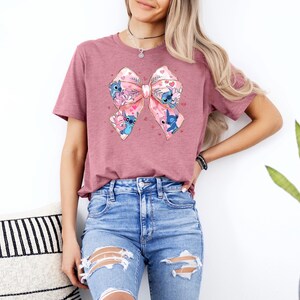 May include: A mauve t-shirt with a pink bow featuring multiple blue Stitch characters from the Disney movie Lilo & Stitch. The bow has white hearts and pink and white polka dots.