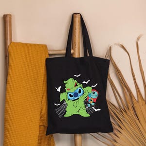 May include: Black tote bag featuring a green and blue cartoon character dressed as a Halloween monster, with white bats and a spider web design. The bag is hanging on a wooden rack with a yellow blanket.