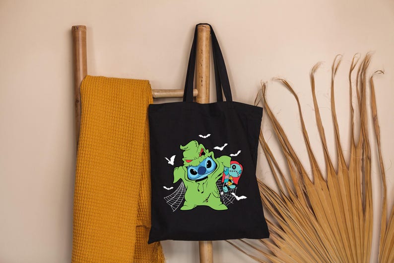 May include: Black tote bag featuring a green and blue cartoon character dressed as a Halloween monster, with white bats and a spider web design. The bag is hanging on a wooden rack with a yellow blanket.