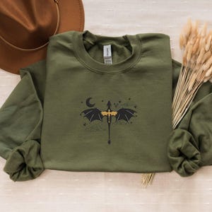 May include: A green long-sleeved sweatshirt featuring a black and gold dragon graphic. The dragon is flying with its wings spread and a crescent moon behind it.
