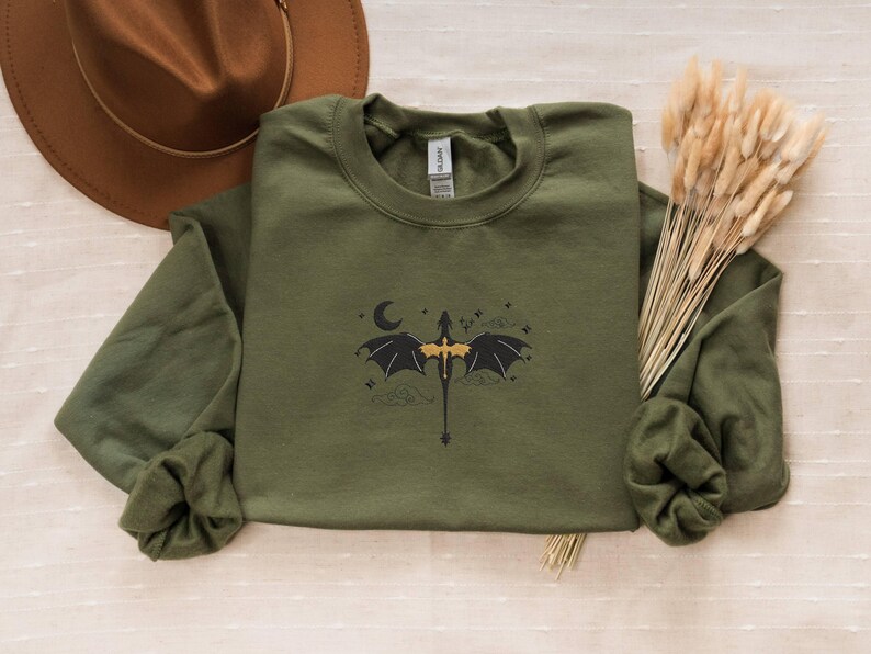 May include: A green long-sleeved sweatshirt featuring a black and gold dragon graphic. The dragon is flying with its wings spread and a crescent moon behind it.