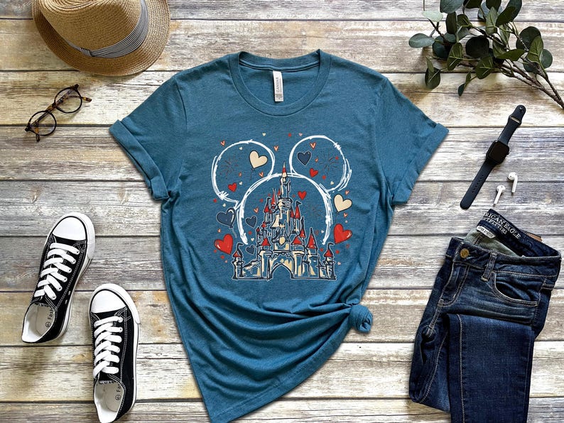 May include: Teal t-shirt featuring a castle design within Mickey Mouse ears, surrounded by hearts. The shirt is paired with black sneakers, jeans, and accessories. A casual, themed apparel item.