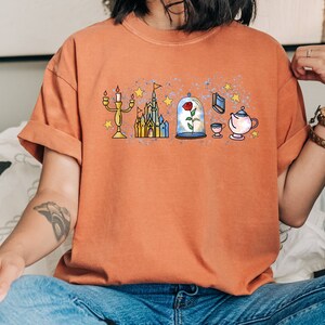 Puede incluir: Una mujer lleva una camiseta naranja quemada con un dise&ntilde;o gr&aacute;fico de un castillo, una rosa en una c&uacute;pula de cristal, una tetera, una taza y un candelabro. El dise&ntilde;o es de estilo de dibujos animados con un efecto acuarela.