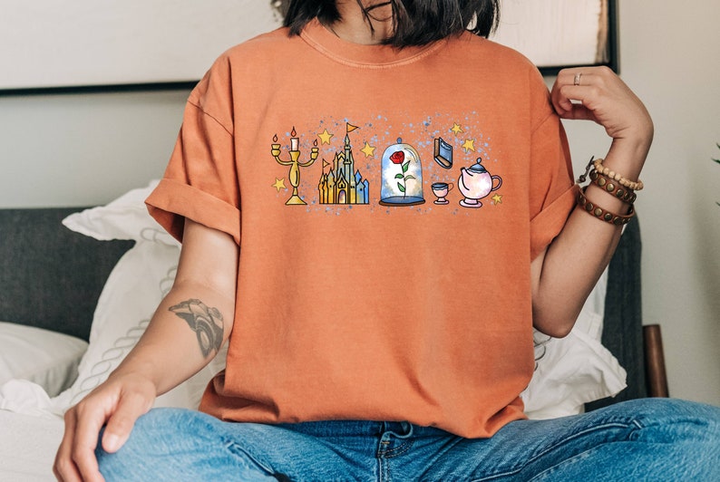 Puede incluir: Una mujer lleva una camiseta naranja quemada con un dise&ntilde;o gr&aacute;fico de un castillo, una rosa en una c&uacute;pula de cristal, una tetera, una taza y un candelabro. El dise&ntilde;o es de estilo de dibujos animados con un efecto acuarela.