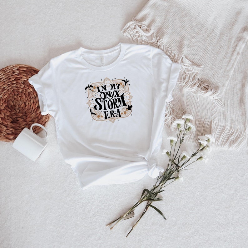 May include: White t-shirt with the text "IN MY ONYX STORM ERA" in a decorative design. The design includes bats and stars in black and tan colors. The shirt is styled on a white surface with a woven basket and a white mug.