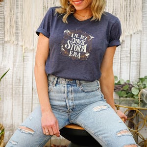 May include: A navy blue t-shirt with rolled sleeves featuring the text "IN MY ONYX STORM ERA" in a decorative design. The shirt is paired with light blue ripped jeans. The overall aesthetic is casual and stylish.
