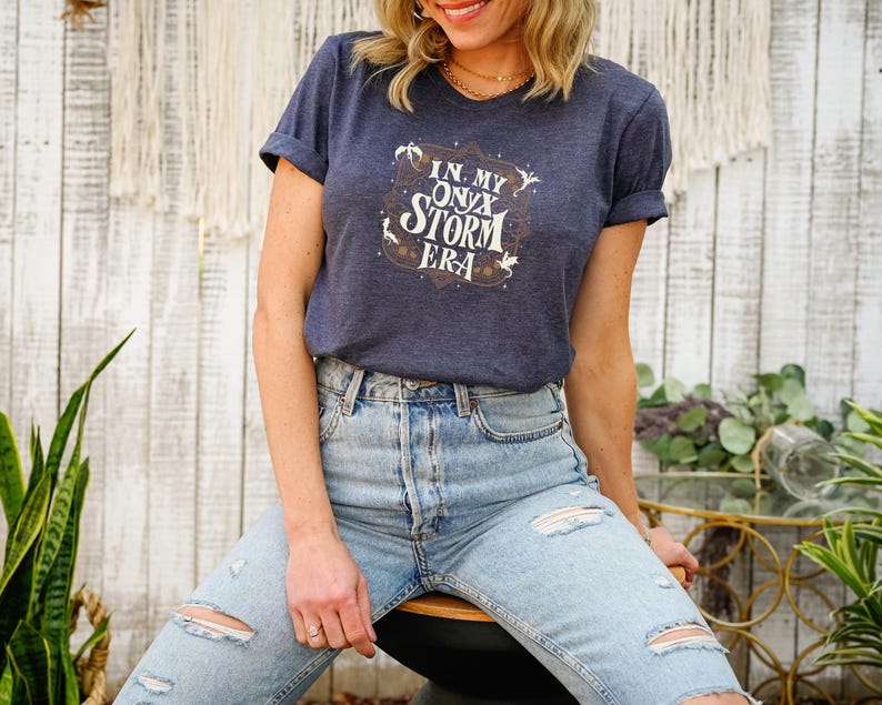 May include: A navy blue t-shirt with rolled sleeves featuring the text "IN MY ONYX STORM ERA" in a decorative design. The shirt is paired with light blue ripped jeans. The overall aesthetic is casual and stylish.