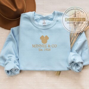 May include: A light blue sweatshirt with gold embroidered text and a Mickey Mouse head design. The text reads "Minnie & Co. Est. 1928". A brown hat is visible in the background. The sweatshirt is a personalized gift.
