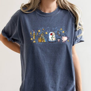 Puede incluir: Una camiseta azul oscuro con un estampado gr&aacute;fico de un castillo, una rosa en una c&uacute;pula de cristal, una tetera, una taza y un candelabro. El gr&aacute;fico est&aacute; rodeado de estrellas.