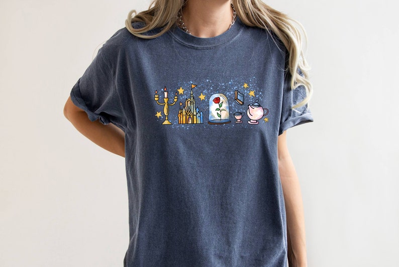 Puede incluir: Una camiseta azul oscuro con un estampado gr&aacute;fico de un castillo, una rosa en una c&uacute;pula de cristal, una tetera, una taza y un candelabro. El gr&aacute;fico est&aacute; rodeado de estrellas.