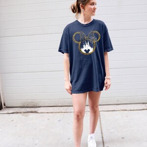 May include: A navy blue t-shirt with a gold glitter Mickey Mouse head design featuring a white silhouette of Cinderella's castle inside the ears. The design also includes a gold glitter bow and fireworks.