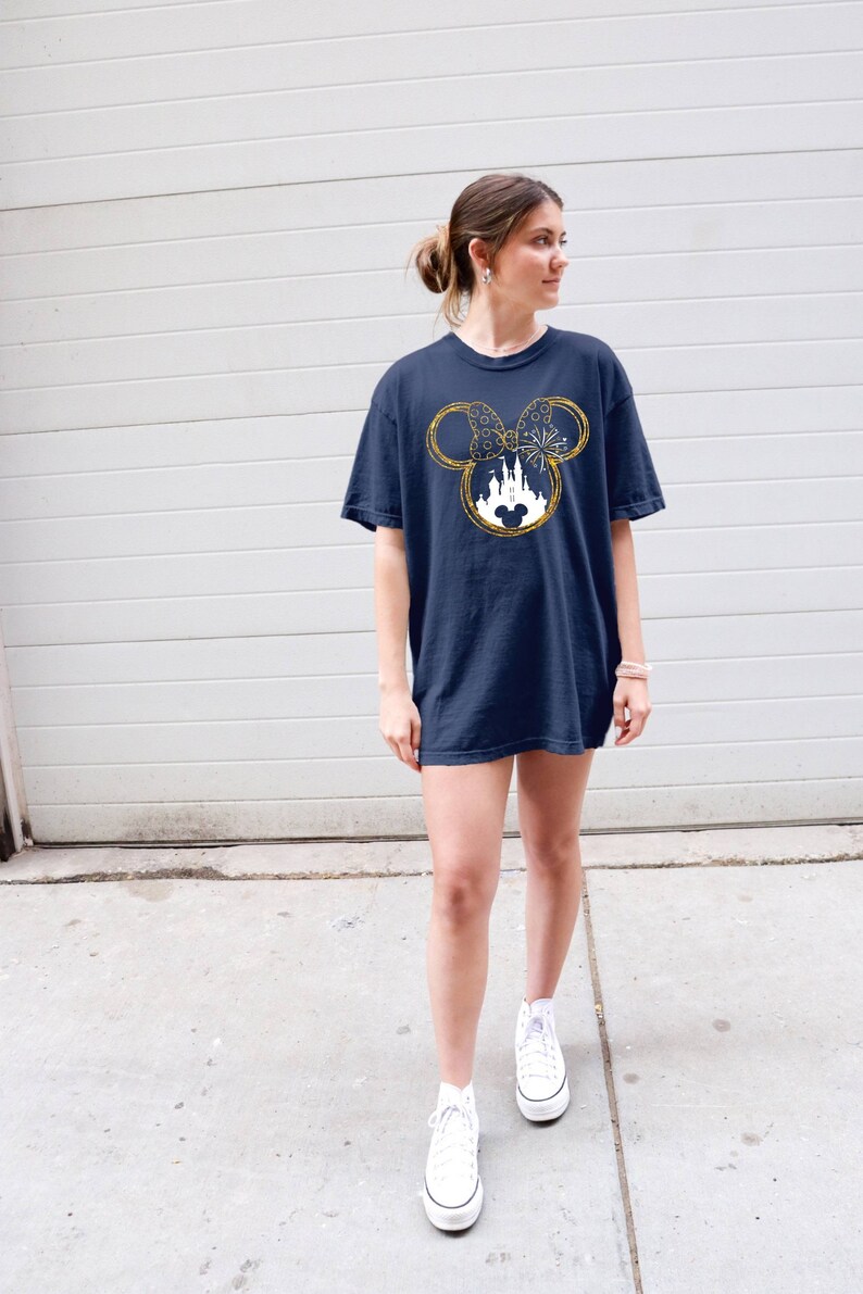 May include: A navy blue t-shirt with a gold glitter Mickey Mouse head design featuring a white silhouette of Cinderella's castle inside the ears. The design also includes a gold glitter bow and fireworks.