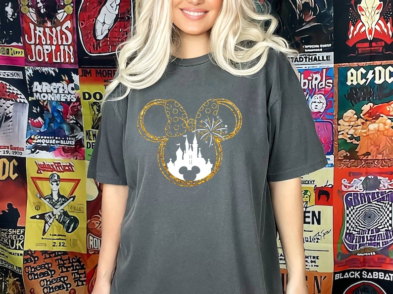 May include: A charcoal grey t-shirt with a gold glitter Mickey Mouse silhouette. The silhouette features a white castle and fireworks inside the ears.