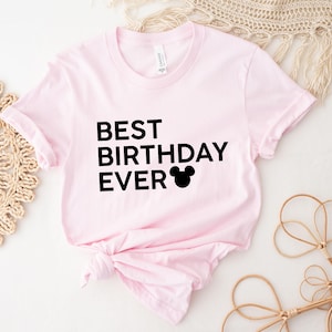 Peut inclure: T-shirt rose avec un texte noir qui dit "Meilleur anniversaire de tous les temps" et une silhouette noire de Mickey Mouse.