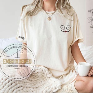 K&ouml;nnte beinhalten: Cremefarbenes T-Shirt mit einem Sally-Gesicht-Design und dem Text "His Sally" auf dem &Auml;rmel. Das Shirt hat auch den Text "Personalized Gift" und "Embroidered Comfort Colors" in einem kreisf&ouml;rmigen Logo.