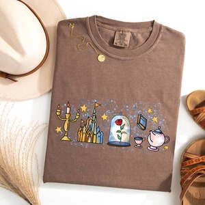 Puede incluir: Una camiseta marr&oacute;n con un estampado gr&aacute;fico de un castillo, una rosa en una c&uacute;pula de cristal, un candelabro, una tetera y una taza. El gr&aacute;fico est&aacute; rodeado de estrellas azules y blancas.