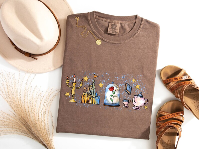 Puede incluir: Una camiseta marr&oacute;n con un estampado gr&aacute;fico de un castillo, una rosa en una c&uacute;pula de cristal, un candelabro, una tetera y una taza. El gr&aacute;fico est&aacute; rodeado de estrellas azules y blancas.