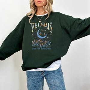 May include: Dark green sweatshirt with the word "VELARIS" in a stylized font above a crescent moon and stars graphic. Below the graphic is the text "CITY OF STARLIGHT". The sweatshirt is a relaxed fit.