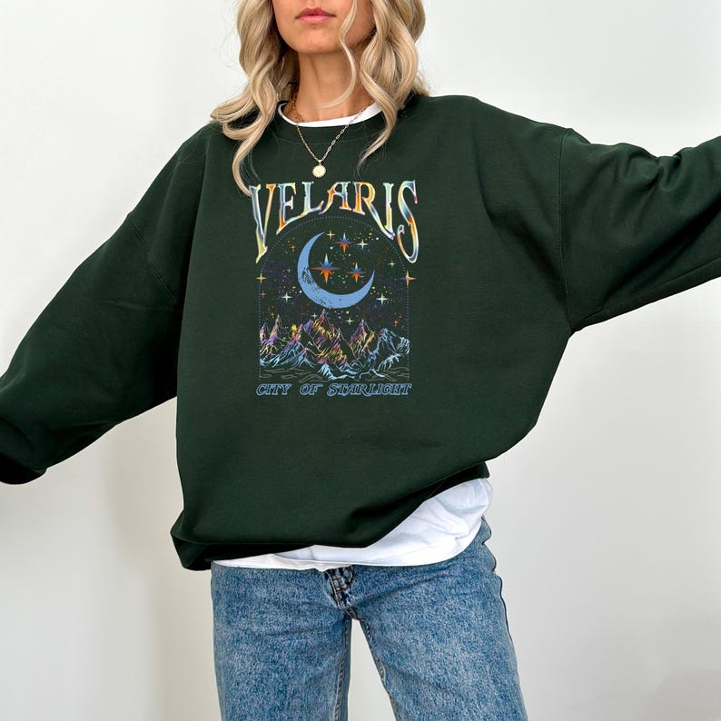 May include: Dark green sweatshirt with the word "VELARIS" in a stylized font above a crescent moon and stars graphic. Below the graphic is the text "CITY OF STARLIGHT". The sweatshirt is a relaxed fit.