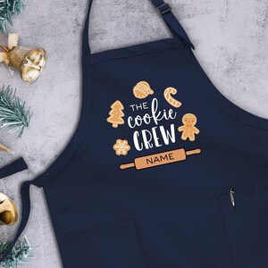 May include: A navy blue apron with a pocket and adjustable straps. It features a white design with the text "The cookie crew" and a rolling pin with the word "NAME" on it. The design also includes gingerbread cookies, a candy cane, a snowflake, and a Christmas tree.