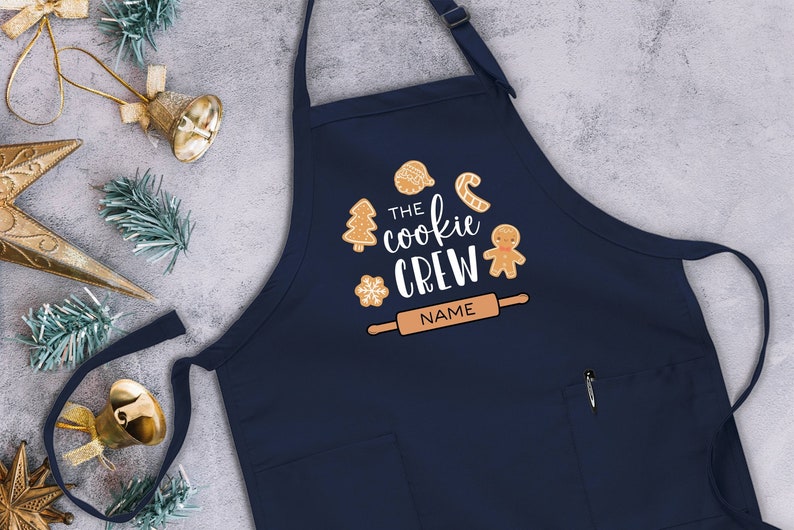May include: A navy blue apron with a pocket and adjustable straps. It features a white design with the text "The cookie crew" and a rolling pin with the word "NAME" on it. The design also includes gingerbread cookies, a candy cane, a snowflake, and a Christmas tree.