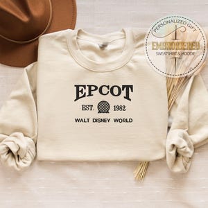 Puede incluir: Sudadera color crema con la palabra "EPCOT" en negro, junto con "EST. 1982" y "WALT DISNEY WORLD". Un sombrero marr&oacute;n es visible en la esquina superior izquierda. La sudadera tiene mangas largas y cuello redondo. Una pegatina circular dice "PERSONALIZED GIFT EMBROIDERED SWEATSHIRT & HOODIE."