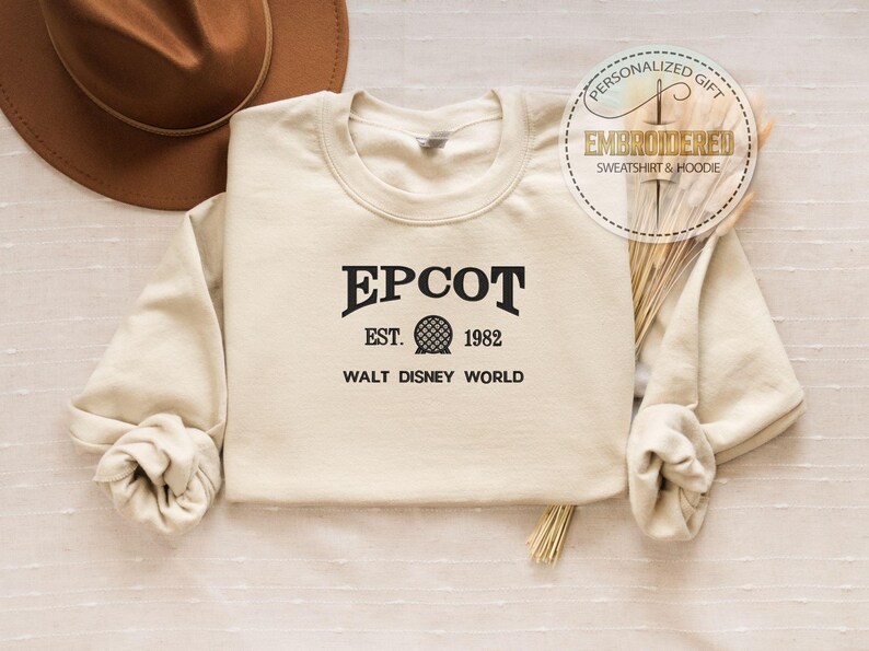 Puede incluir: Sudadera color crema con la palabra "EPCOT" en negro, junto con "EST. 1982" y "WALT DISNEY WORLD". Un sombrero marr&oacute;n es visible en la esquina superior izquierda. La sudadera tiene mangas largas y cuello redondo. Una pegatina circular dice "PERSONALIZED GIFT EMBROIDERED SWEATSHIRT & HOODIE."