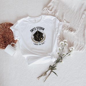 May include: White t-shirt with a black and gold graphic design. The graphic features a stylized dragon with the text "ONYX STORM" and "BRAVE THE DARK".
