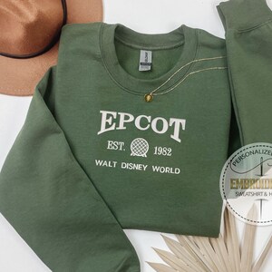 Puede incluir: Sudadera verde EPCOT con letras blancas y un gr&aacute;fico de globo. El texto dice "EPCOT EST. 1982 WALT DISNEY WORLD". Tambi&eacute;n se ven un collar dorado y un sombrero marr&oacute;n.