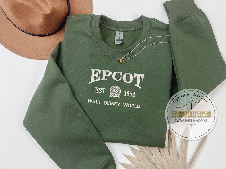 Puede incluir: Sudadera verde EPCOT con letras blancas y un gr&aacute;fico de globo. El texto dice "EPCOT EST. 1982 WALT DISNEY WORLD". Tambi&eacute;n se ven un collar dorado y un sombrero marr&oacute;n.