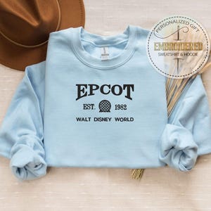 Puede incluir: Una sudadera azul claro de EPCOT con el texto "EPCOT EST. 1982 WALT DISNEY WORLD" en negro. La sudadera tiene mangas largas y cuello redondo. Un sombrero marr&oacute;n es visible en el fondo. Una pegatina circular dice "REGALO PERSONALIZADO SUDADERA Y SUDADERA CON CAPUCHA BORDADA".