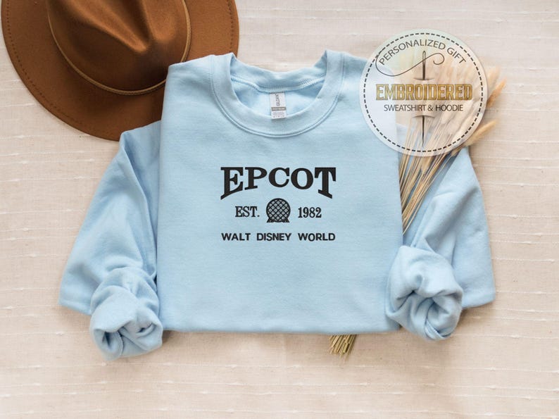 Puede incluir: Una sudadera azul claro de EPCOT con el texto "EPCOT EST. 1982 WALT DISNEY WORLD" en negro. La sudadera tiene mangas largas y cuello redondo. Un sombrero marr&oacute;n es visible en el fondo. Una pegatina circular dice "REGALO PERSONALIZADO SUDADERA Y SUDADERA CON CAPUCHA BORDADA".