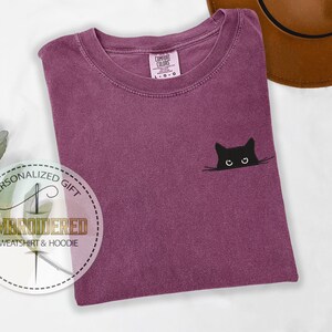 May include: A purple t-shirt with a black cat peeking out from behind a white line. The cat has white eyes and a black nose. The t-shirt has the text "COMFORT COLORS" on the tag.