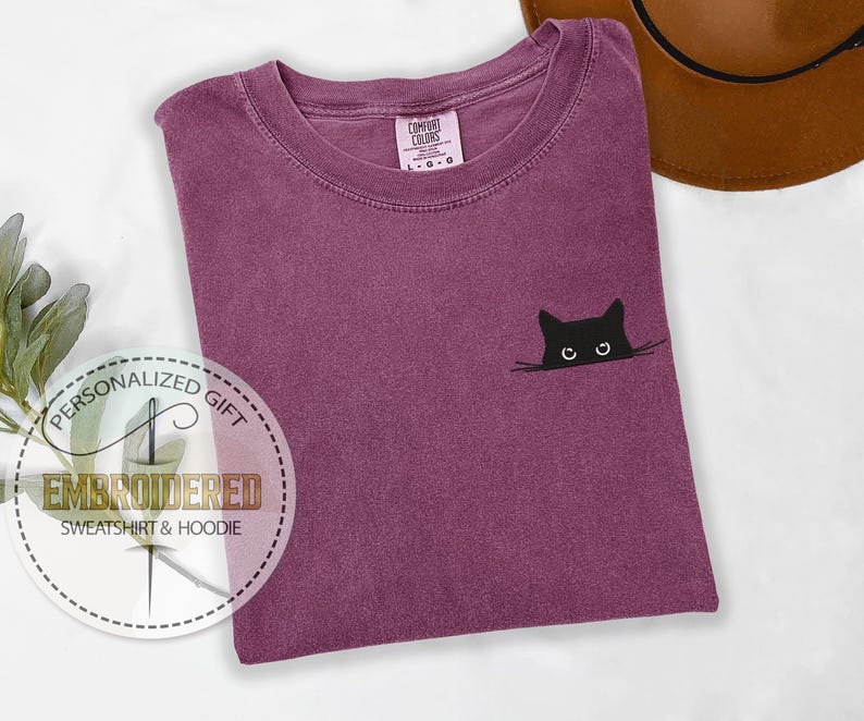 May include: A purple t-shirt with a black cat peeking out from behind a white line. The cat has white eyes and a black nose. The t-shirt has the text "COMFORT COLORS" on the tag.