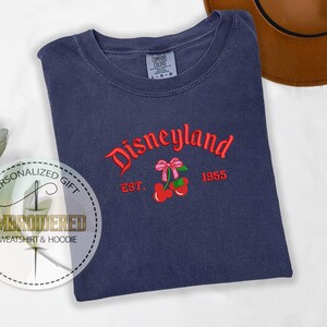 Op de afbeelding: Een marineblauw T-shirt met de geborduurde tekst "Disneyland Est. 1955" in rood en wit. De borduurwerk bevat ook een rode kers met een roze strik.
