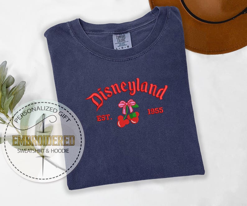 Op de afbeelding: Een marineblauw T-shirt met de geborduurde tekst "Disneyland Est. 1955" in rood en wit. De borduurwerk bevat ook een rode kers met een roze strik.