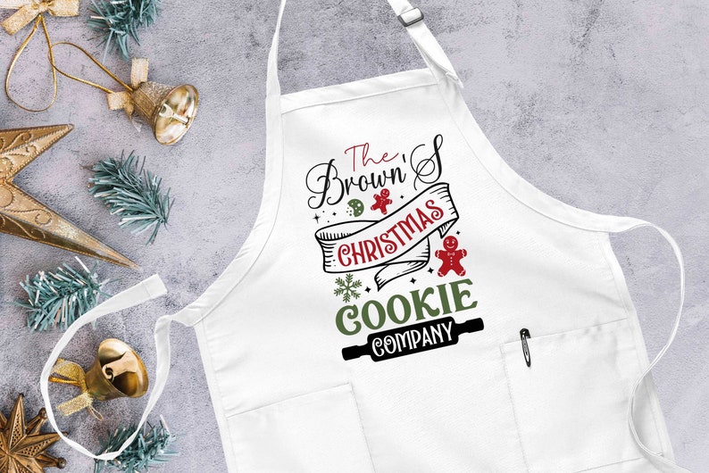Puede incluir: Delantal blanco con un dise&ntilde;o rojo y verde que dice "The Brown's Christmas Cookie Company" con un hombre de jengibre y un rodillo.
