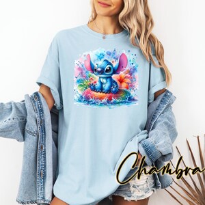 Może przedstawiać: Jasnoniebieska koszulka z akwarelową ilustracją Stitcha z Lilo & Stitch. Grafika przedstawia Stitcha siedzącego na tęczowej powierzchni otoczonego kolorowymi kwiatami i rozpryskami wody. Koszulka ma kr&oacute;tkie rękawy.