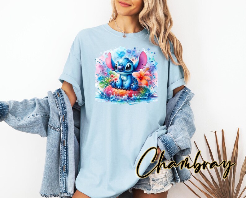 Może przedstawiać: Jasnoniebieska koszulka z akwarelową ilustracją Stitcha z Lilo & Stitch. Grafika przedstawia Stitcha siedzącego na tęczowej powierzchni otoczonego kolorowymi kwiatami i rozpryskami wody. Koszulka ma kr&oacute;tkie rękawy.