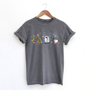 Puede incluir: Una camiseta gris oscuro con un estampado gr&aacute;fico de objetos ic&oacute;nicos de la pel&iacute;cula de Disney La Bella y la Bestia. El gr&aacute;fico incluye un candelabro, un castillo, una rosa en una c&uacute;pula de vidrio, un libro y una tetera.