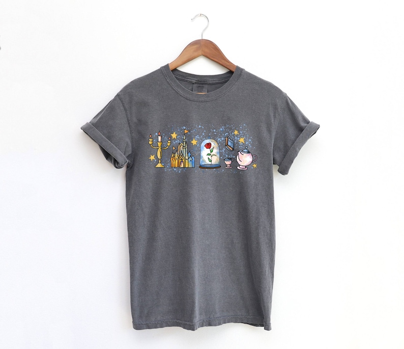 Puede incluir: Una camiseta gris oscuro con un estampado gr&aacute;fico de objetos ic&oacute;nicos de la pel&iacute;cula de Disney La Bella y la Bestia. El gr&aacute;fico incluye un candelabro, un castillo, una rosa en una c&uacute;pula de vidrio, un libro y una tetera.