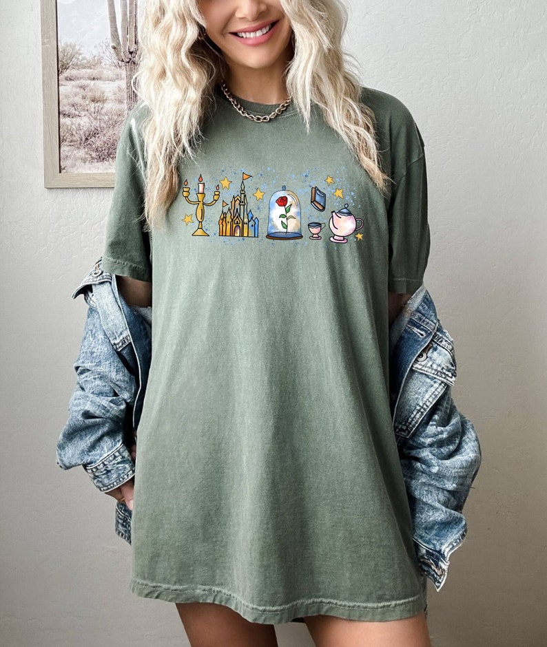 Puede incluir: Una camiseta verde con un estampado gr&aacute;fico de un castillo, una rosa en una c&uacute;pula de cristal, un candelabro, un libro y una tetera. El gr&aacute;fico est&aacute; en estilo de dibujos animados y est&aacute; rodeado de estrellas.