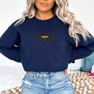 May include: A navy blue sweatshirt featuring a black dragon graphic with outstretched wings and a crescent moon. The dragon is gold in colour.